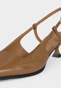 Γυαλιστερό καφέ παπούτσι slingback με μυτερή μύτη, λεπτό λουράκι στον αστράγαλο με χρυσή αγκράφα και χαμηλό τακούνι kitten σε ανοιχτό φόντο.