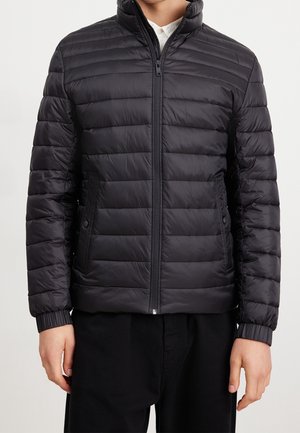 Homme portant une doudoune matelassée noire avec une fermeture éclair à l’avant et deux poches latérales sur une chemise blanche.