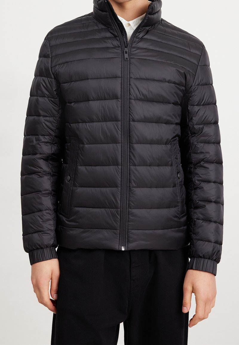 Homme portant une doudoune matelassée noire avec une fermeture éclair à l’avant et deux poches latérales sur une chemise blanche.
