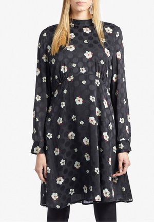 Kvinde iført en sort langærmet kjole med hvidt blomsterprint og diskret prikket mønster, stående med armene afslappet langs siderne.