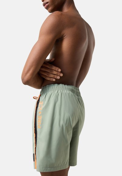 Homme torse nu, bras croisés, portant un short de bain vert clair avec le texte orange "FIRE + ICE" sur le côté et une ceinture élastique.