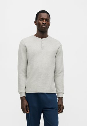 Mees, kellel on seljas helehall pika varrukaga henley-särk, millel on kolm nööpi, ja tumesinised püksid, seisab ühevärvilise tausta ees.