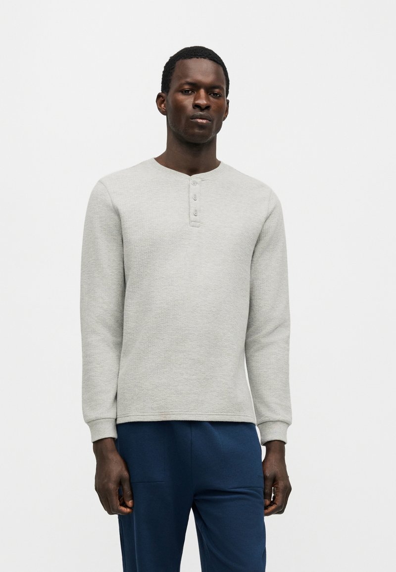 Άνδρας που φοράει ανοιχτό γκρι μακρυμάνικο πουκάμισο Henley με τρία κουμπιά και σκούρο μπλε παντελόνι, στέκεται μπροστά σε απλό φόντο.