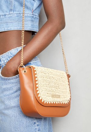 Frau mit dunkler Haut trägt Denim-Shorts und ein Cropped-Top und hält eine hellbraune Lederschultertasche mit geflochtener Klappe und goldener Kettenriemen.