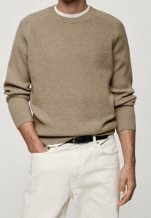 Mand iført en beige strikket sweater over en hvid skjorte, der er stoppet ned i hvide bukser med et sort bælte, den ene hånd i lommen og den anden afslappet.
