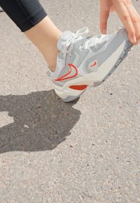 Grijze sportieve schoen met rode accenten, textuurmateriaal, vetersluiting en een dikke witte zool, geplaatst op een betonnen oppervlak.