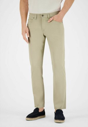 Mann trägt beige, eng anliegende Hosen mit Seitentaschen und schwarze Loafer, steht vor einem einfarbigen Hintergrund.