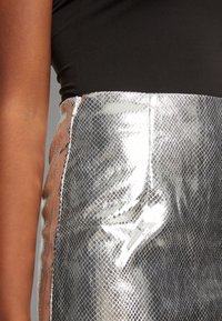Falda de malla plateada con una blusa negra, que presenta una superficie brillante y texturizada, abertura lateral y detalles cosidos a lo largo de los bordes.