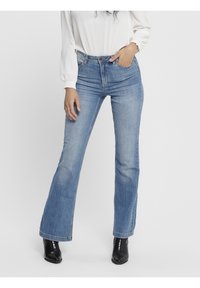 Nainen, jolla on päällään vaaleansiniset bootcut-farkut, valkoinen pitkähihaisia paita ja mustat nilkkurit, yksi käsi taskussa.