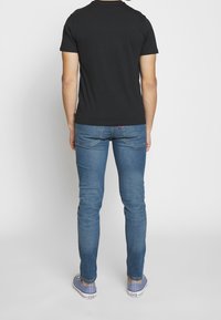 Schwarzes Baumwoll-T-Shirt, getragen mit blauen Denim-Jeans. Die Passform ist körpernah, mit sichtbaren Nähten und dezenten Logo-Akzenten an den Jeans.