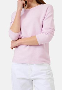 Kvinna som bär en ljusrosa långärmad tröja med uppvikta ärmslut och vita byxor, poserande med en arm i kors och handen nära hakan.