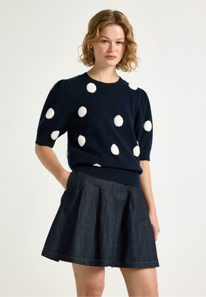 Kvinde iført en navyblå sweater med hvide pomponer og et mørkt plisseret denimskørt, stående med den ene hånd i lommen.