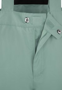 Hellgrüne wasserdichte Hose mit glatter Textur, ausgestattet mit einem vorderen Reißverschluss, Druckknopfverschluss und verstellbaren Trägern.