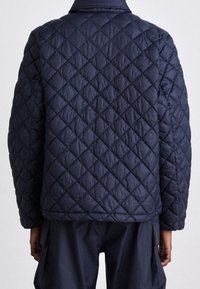 Giacca trapuntata blu navy con motivo a rombi, materiale leggero, colletto alto e spalle scese. La vista dorsale mostra la texture liscia e le cuciture.