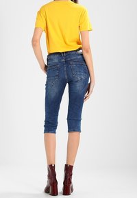 Mujer vestida con una camiseta amarilla de manga corta, jeans azules cortos y botas de tacón rojas, de pie con la espalda hacia la cámara.