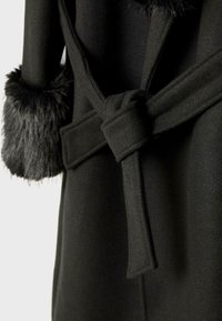 Sort uldfrakke med bundet bælte og sorte faux fur-manchetter på ærmerne, præsenteret mod en ensfarvet lys baggrund.