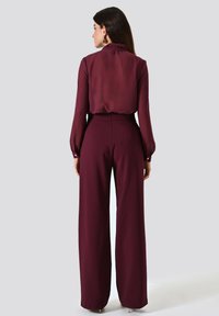 Blouse bordeaux à col haut et manches longues, associée à un pantalon bordeaux taille haute et à jambes larges. Texture lisse, détails minimalistes.
