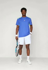 Sininen urheilu t-paita vihreillä yksityiskohdilla, yhdistettynä valkoisiin shortsiesi. Pelaaja pitää tennismailaa, ja hänen jalassaan on valkoiset lenkkitossut sekä rannekkeet.