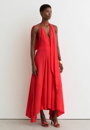 Kobieta w zwiewnej, asymetrycznej czerwonej sukience z głębokim dekoltem w kształcie litery V oraz pasujących czerwonych sandałach na wysokim obcasie na jednolitym białym tle.