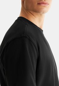 T-shirt noir à manches longues avec un col rond, au tissu doux et à la couture visible sur les coutures des épaules. Coupe décontractée, vue de côté.