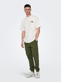 Camiseta blanca de algodón con un logo en el pecho, combinada con pantalones cargo verdes y zapatillas blancas. Estilo casual, ajuste relajado con bolsillos visibles.