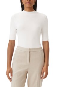 Mujer con una blusa blanca ajustada de manga corta y cuello alto, vestida con pantalones beige a medida, de pie con los brazos relajados a los lados.