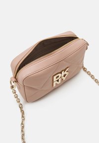 Petit sac bandoulière en cuir beige avec fermeture éclair dorée et bandoulière en chaîne, lettres du logo dorées à l'avant "DKNY," et ouverture supérieure montrant l'intérieur.