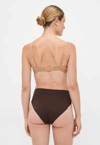 Woman with braided bun hairstyle wearing a beige bra and high-waisted dark brown underwear, standing facing away against a plain white background.