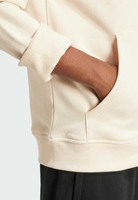 Beige pullover hoodie with a textured cuff detail, featuring a side pocket and a relaxed fit. The fabric appears soft and warm.