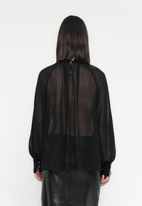 Donna con capelli scuri e lisci, che indossa una blusa nera trasparente a maniche lunghe e una gonna di pelle nera, vista di schiena su uno sfondo bianco.
