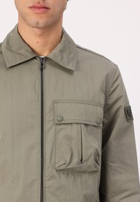 Jachetă de culoare verde măslinie deschisă, cu fermoar frontal, guler ascuțit și două buzunare frontale. Fabricată dintr-un material neted, cu un patch cu logo pe mânecă.