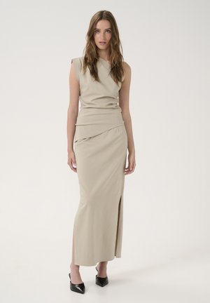 Beige sleeveless dress with draped design, side slit, and smooth texture, paired with pointed black shoes. Elegant and fitted silhouette.