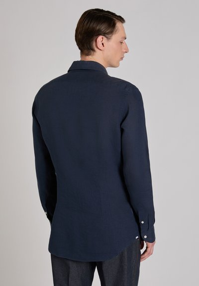 Hombre de perfil, vistiendo una camisa de manga larga azul marino oscuro y pantalones oscuros, frente a un fondo liso y claro.