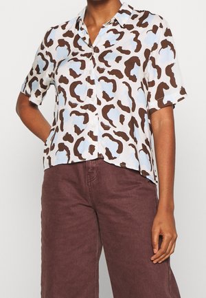 Korte mouwen overhemd met knoopsluiting met bruin en lichtblauw abstract dessin, gedragen met hoge taille bruine broek door een staand persoon.