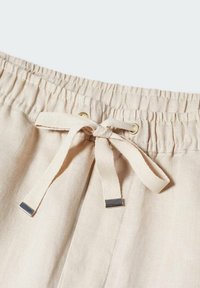 Gros plan sur un pantalon en tissu beige avec taille élastique et cordon noué avec embouts métalliques et œillets dorés.