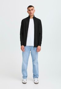Svart zip-up jakke med høy krage, sammen med en hvit T-skjorte og lyseblå jeans. Modellen står i uformelle sko mot en ensfarget bakgrunn.