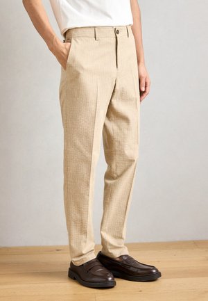 Beige gemusterte Hose mit geradem Schnitt, ausgestattet mit Seitentaschen und einem Knopfverschluss, kombiniert mit dunkelbraunen Slippern auf einem Holzboden.