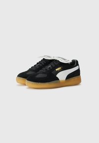 Sorte sømdækkede sneakers med en hvid læderstribe, gyldent logo og en gennemsigtig gummisål. Rund tå og flade snørebånd.
