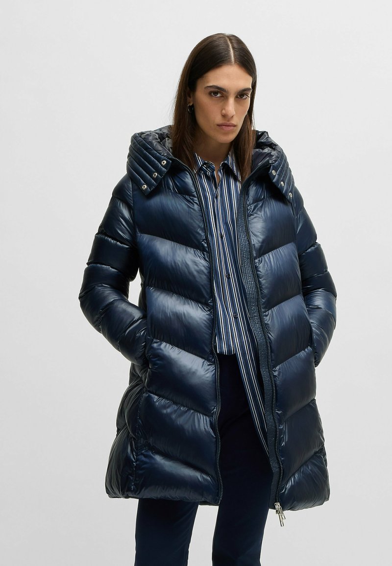 Kobieta ubrana w błyszczący granatowy puchowy płaszcz z kapturem, pod który ma na sobie niebieską koszulę w paski i ciemne spodnie, stoi na tle jasnej, jednolitej ściany.