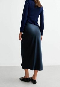 Marineblaues Langarmshirt aus strukturiertem Stoff kombiniert mit einem dunklen Satinhrock in Midi-Länge. Das Outfit wird von schwarzen flachen Schuhen mit dekorativen Akzenten ergänzt.