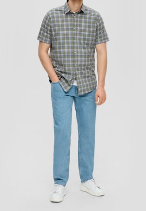 Mężczyzna w zielono-fioletowej kraciastej koszuli z krótkim rękawem, jasnoniebieskich dżinsach i białych trampkach, stojący z jedną ręką w kieszeni.