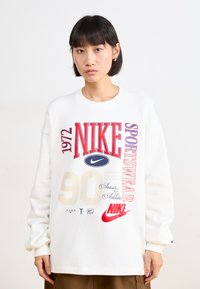 Balta sporta krekla ar drosmīgu sarkanu un zilu "NIKE" tekstu, gada "1972", un grafiskajiem elementiem, tostarp "90" un logotipiem. Brīvs piegriezums ar garām piedurknēm.