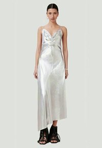 Mulher vestindo um vestido de seda satinada prateada com frente franzida e barra assimétrica, combinado com sandálias pretas de salto aberto com franjas, posando contra um fundo liso.