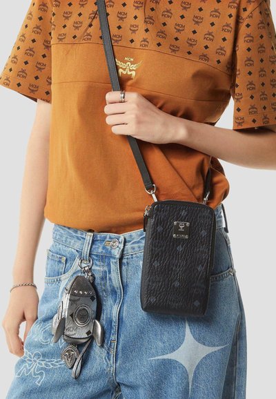 Persoon draagt een bruin MCM-logo shirt en een blauwe spijkerbroek met een zilveren raket sleutelhanger en een kleine zwarte, gestructureerde crossbodytas.