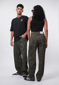 Green leopard-print pants with a loose fit, paired with a black t-shirt. The pants feature a back label and side pockets. Black shoes complete the look.