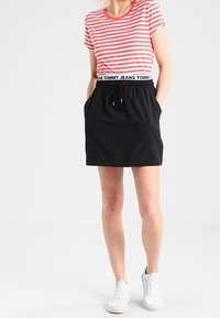Rot und weiß gestreiftes Kurzarmhemd kombiniert mit einem schwarzen Rock mit elastischem Bund und zwei Seitentaschen. Das Model trägt weiße Sneakers.