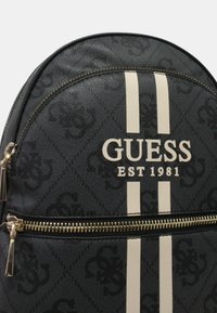 Sac à dos noir avec un logo Guess et des rayures blanches, doté d'une poche zippée à l'avant et d'un motif texturé sur la surface.