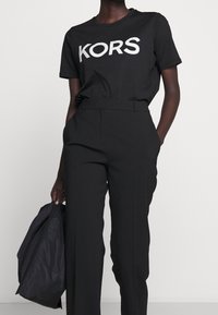 Musta t-paita, jossa hopeinen "KORS"-logo, yhdistetty mustiin räätälöityihin housuihin. Malli pitää mustaa laukkua, joka esittelee yksinkertaista, tyylikästä muotoilua.