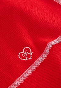 Punane kangas ribast tekstuuriga, millel on valge pitsäär ja kattega südame tikand, mis asub alumises nurgas.