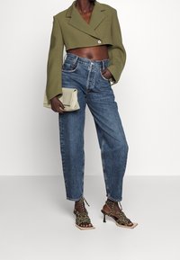Blazer corto verde oliva con maniche ampie, jeans in denim blu a vita alta e sandali con tacco neri con cinturini e dettagli a catena.